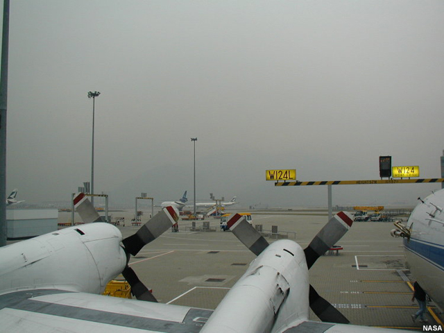 Photo from on top of parked airplane's wing showing foggy/hazy conditions filtering through the airport.